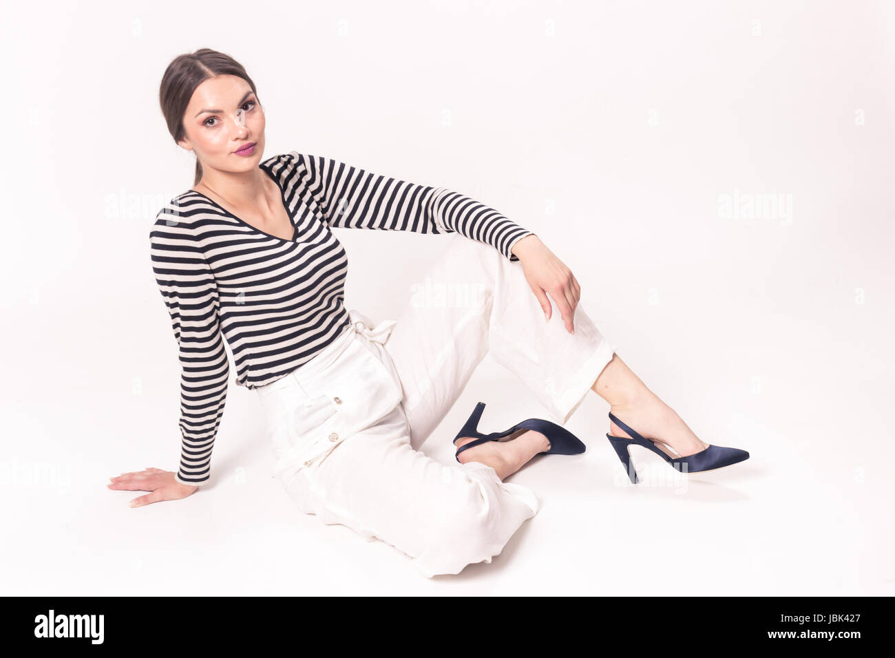 L'un young Caucasian woman 20s, 29 ans, mannequin, posant assis fond blanc, studio, horizontal stripe shirt, pantalon blanc, haut Banque D'Images