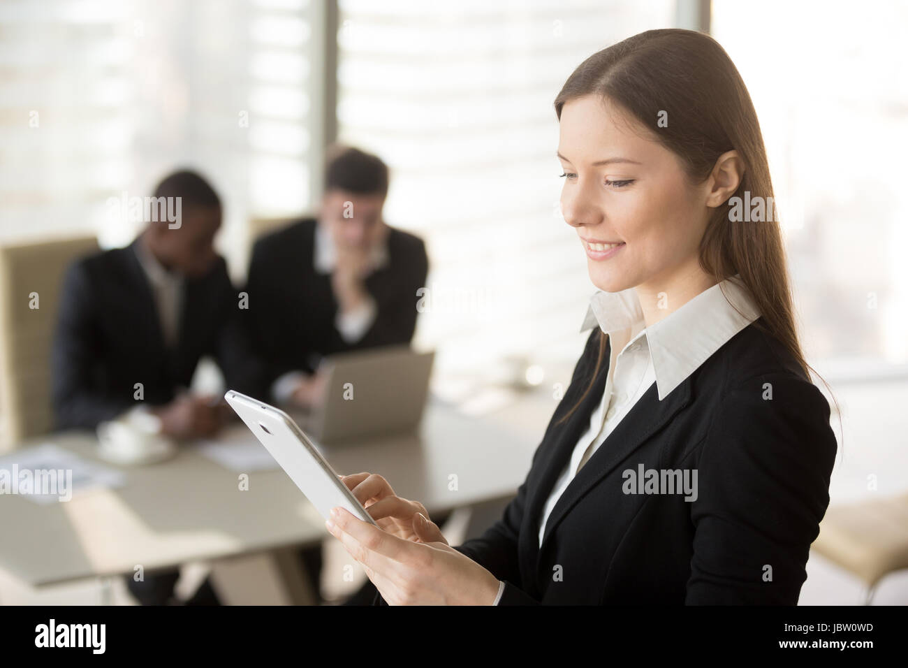 Young attractive businesswoman holding tablet, regardant l'écran, standing in office avec nos collègues de l'arrière-plan, l'organisation de réunion de planification, l'usi Banque D'Images