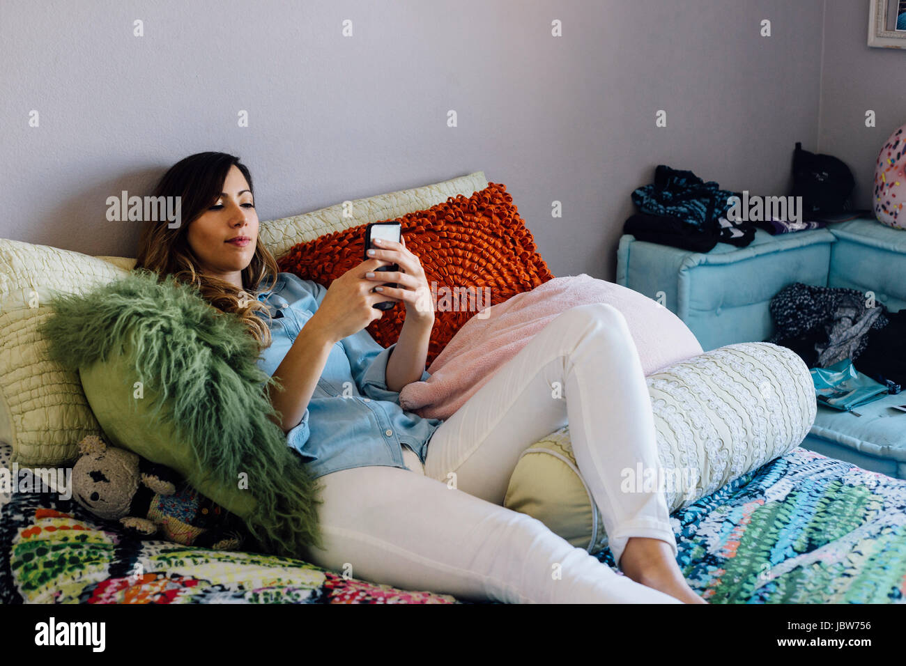 Jeune femme couchée sur un lit shabby chic looking at smartphone Banque D'Images
