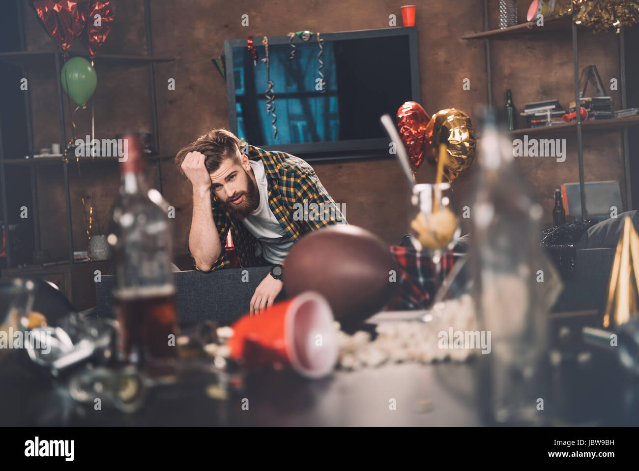 Jeune homme barbu avec gueule assis sur table en salle malpropre after party Banque D'Images