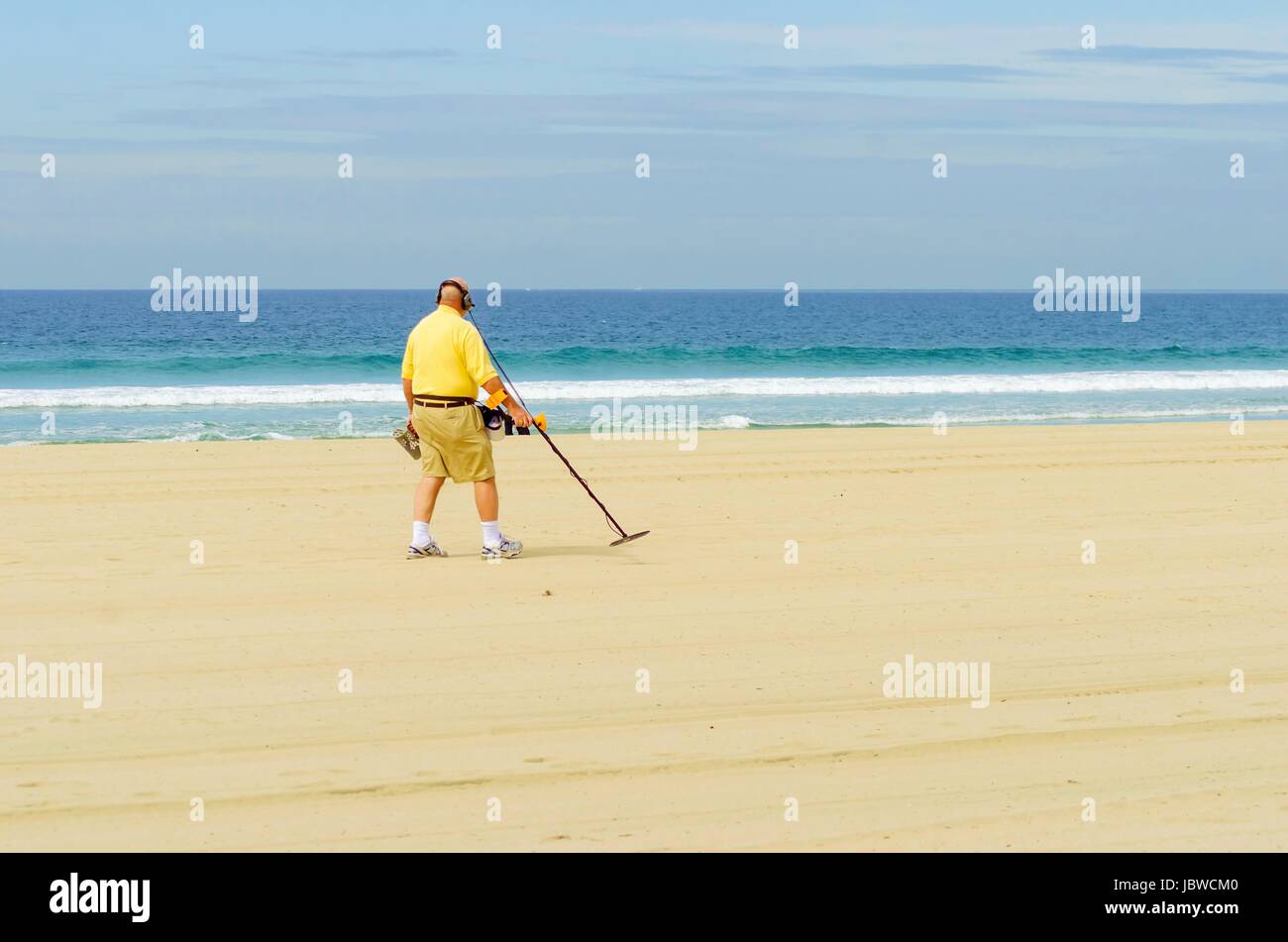 Un vieil homme marchant le long de la côte, la tenue d'un détecteur de métal portable sur la plage du Pacifique en Amérique, en pointant le long bâton au sable, essayant de trouver des objets métalliques et la zone de numérisation portant des écouteurs. Banque D'Images