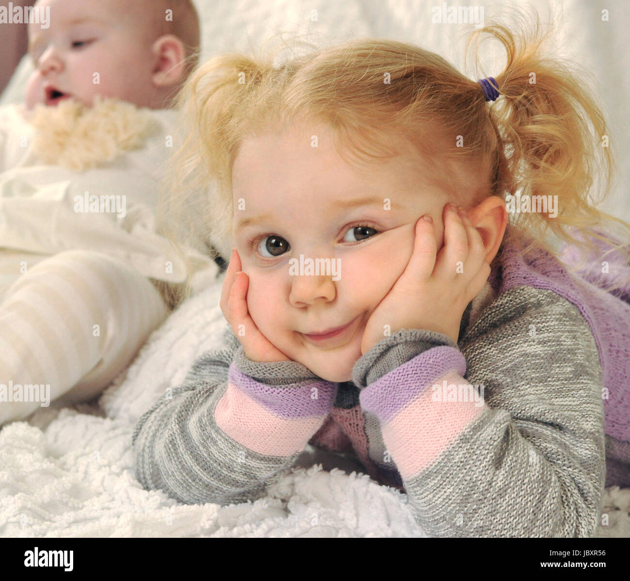 Adorable Petite Fille Blonde Au Lit Sur Le Ventre Se Penchant Sa Tete Dans Ses Mains Tout En Regardant A L Appareil Photo Avec Un Curieux L Ennui Et L Expression Du Visage A Cote