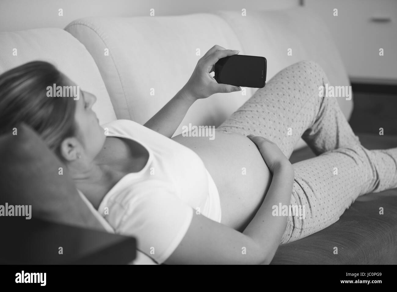 Tourné en noir et blanc gros plan de femme enceinte détente sur canapé et using mobile phone Banque D'Images