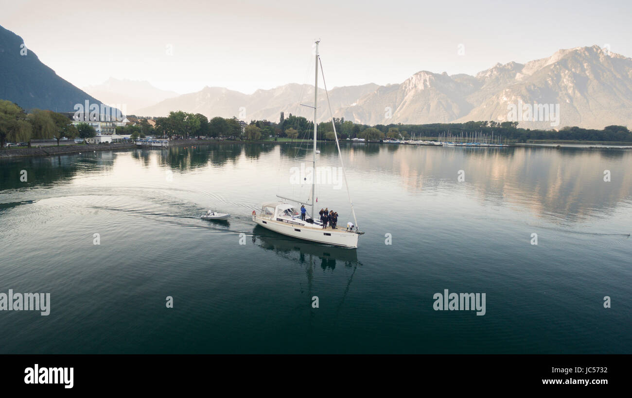 Vue aérienne sur un bateau à voile et traversée en bateau de sauvetage du Lac Léman, avec les montagnes en arrière-plan, des arbres dans les banques du lac et une marina à Villeneuve, Canton de Vaud, Suisse Banque D'Images