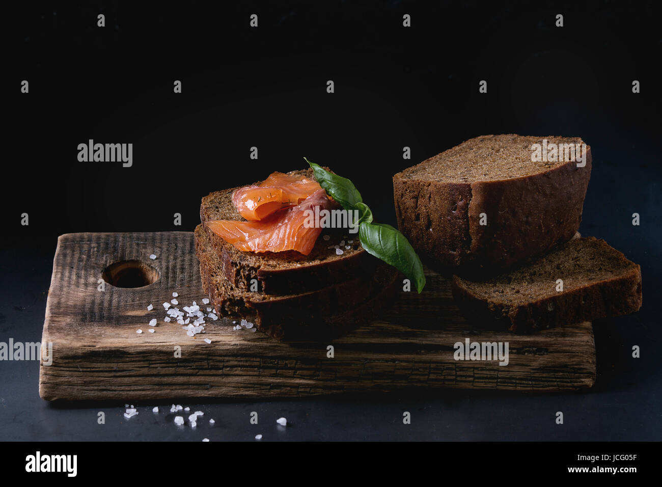 Pile de tranches de pain de seigle fait maison avec du saumon fumé, sel de mer et de basilic frais sur planche en bois sombre sur fond noir. Banque D'Images