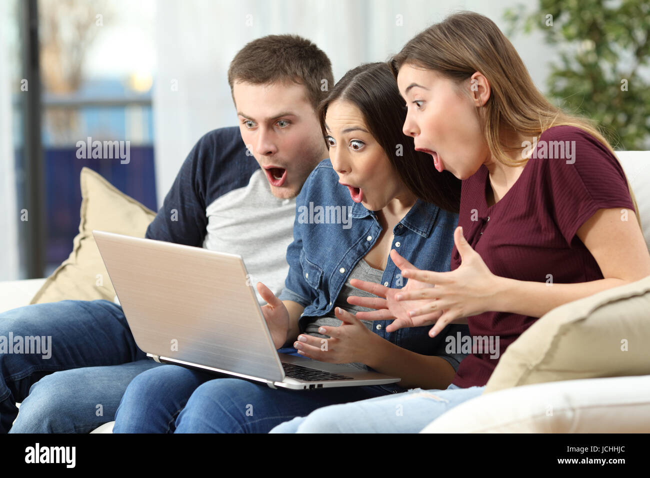 Trois amis surpris à regarder le contenu des médias en ligne en un ordinateur assis sur un canapé dans la salle de séjour à la maison Banque D'Images