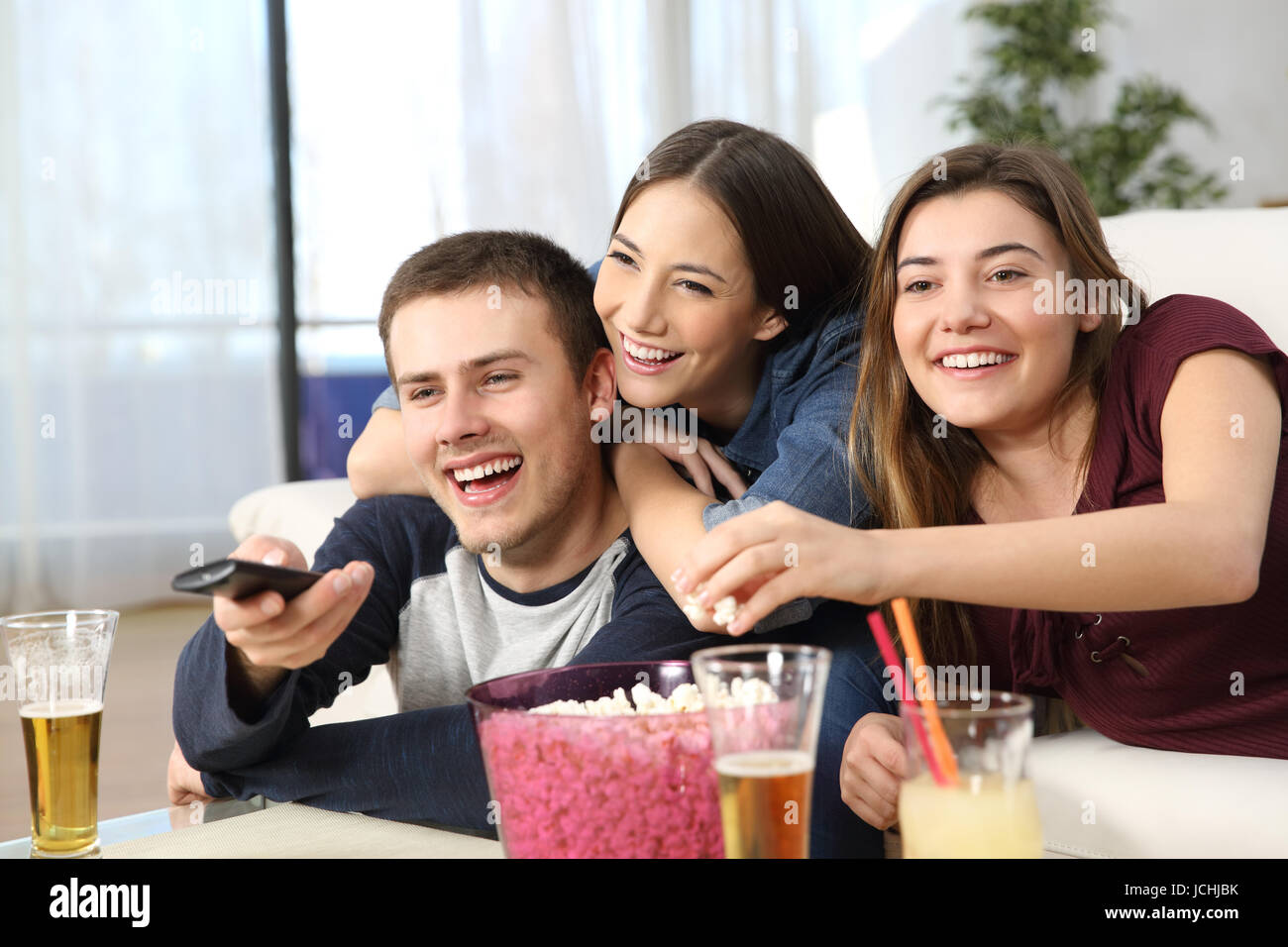 Portrait de trois amis heureux affectueuse watching TV and eating popcorn assis sur un canapé dans la salle de séjour à la maison Banque D'Images