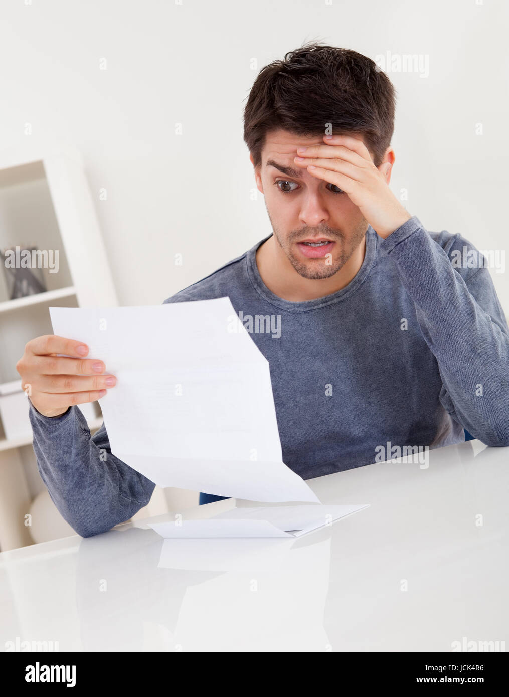 Jeune homme horrifié la lecture d'un document avec une expression de stupeur et sa main à son front comme il fixe de grands yeux à la page de papier Banque D'Images