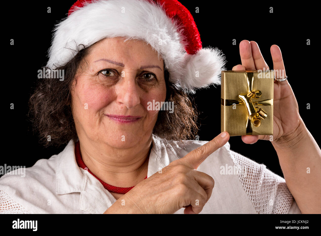 Friendly, vénérable, dark-haired woman wearing un vieux chapeau de Père Noël. Elle est orientée avec son index droit à un petit cadeau de Noël d'or qu'elle tient avec sa main gauche. Banque D'Images