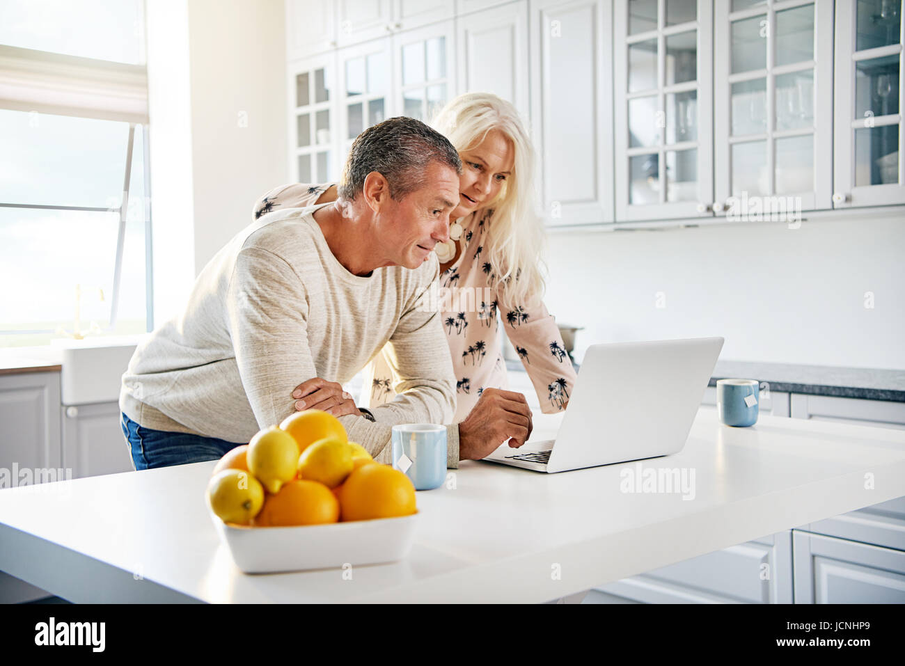 Couple de retraités heureux et en santé la navigation sur internet ainsi que d'un ordinateur portable dans une lumière blanche cuisine avec un bol d'oranges et citrons dans Banque D'Images