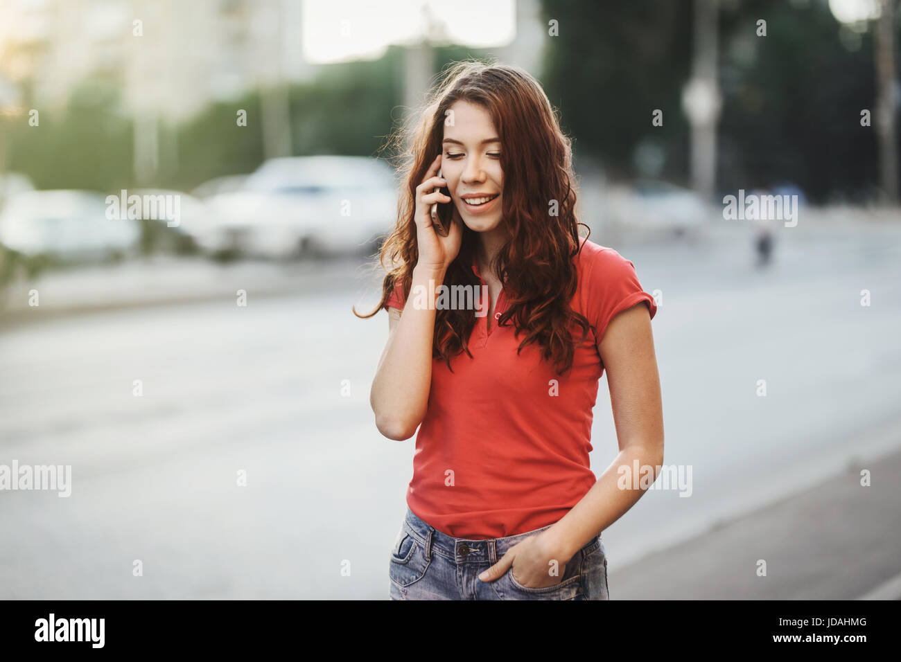 Jolie fille en tenue décontractée est de parler sur un téléphone portable en marche à la ville rue à une heure du coucher de soleil. Banque D'Images