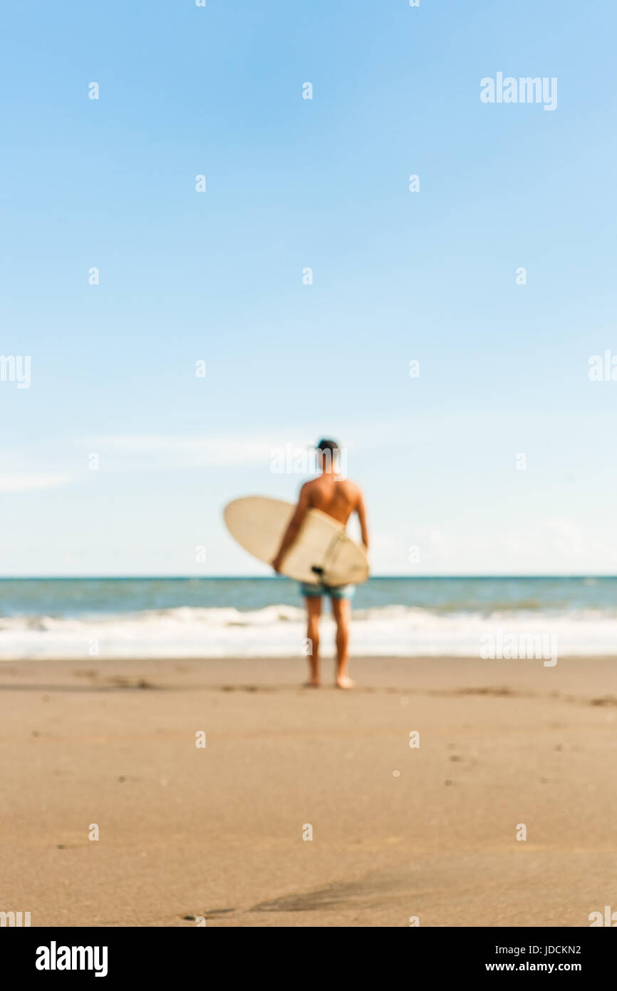 Des problèmes de mise au point floue de bel homme stand avec surf surf long board attendre wave le spot de surf en mer plage de l'océan. Vue depuis l'arrière. Concept o Banque D'Images