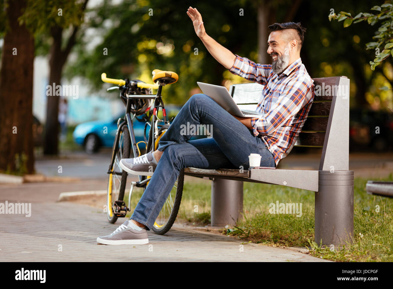 Smiling businessman waving Hello sur une pause-café. Il est assis sur un banc et de travail à l'ordinateur portable, la prochaine audience repose un vélo. Banque D'Images
