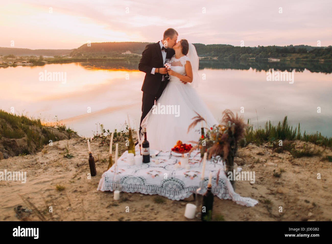 Les nouveaux mariés s'embrasser à l'arrière-plan de la rivière pendant le coucher du soleil derrière l'ensemble de pique-nique Banque D'Images