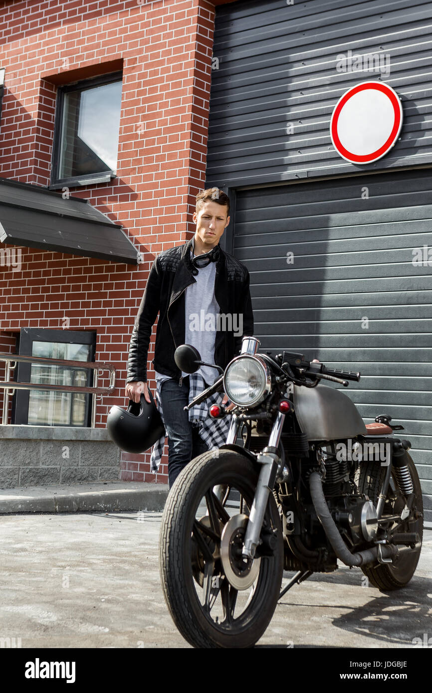 Beau jeune gars en veste biker cuir noir voir son style classique cafe racer motorcycle portes industrielles comme arrière-plan. Fait sur commande de vélo Banque D'Images