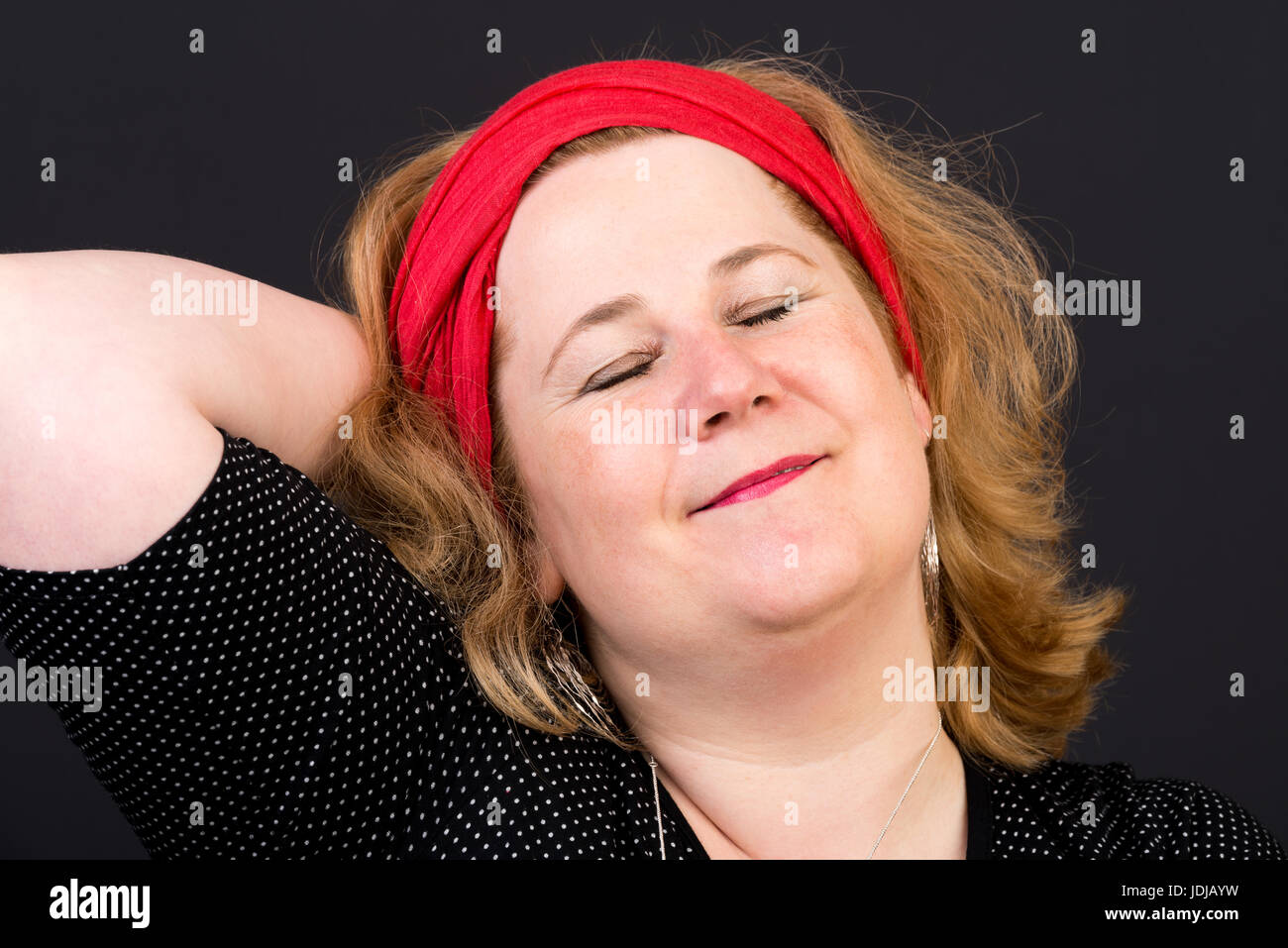 Aux cheveux rouge attrayant embonpoint lite femme mature européenne avec écharpe rouge dans les cheveux avec les yeux fermés, détendue - studio shot sur fond sombre Banque D'Images