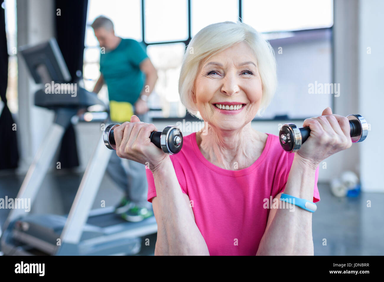 La sportive senior sitting on fitness ball avec haltères, sportif sur tapis roulant derrière dans la haute classe de conditionnement physique Banque D'Images