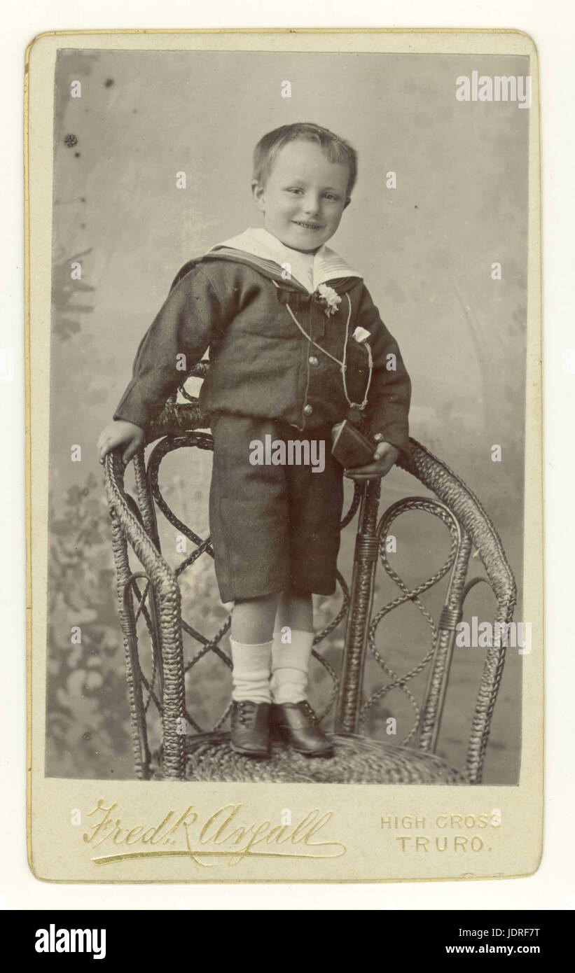 Original CDV (carte de visite) portrait studio de jeune garçon victorien souriant portant un costume de marin tendance debout sur une chaise en osier, toile de fond peinte, Truro, Cornwall, Royaume-Uni vers 1890 Banque D'Images