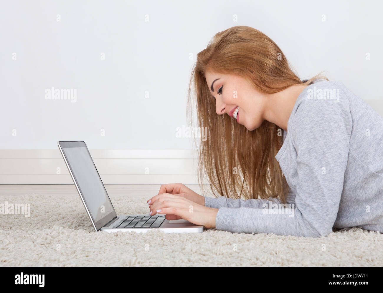 Portrait Of A Smiling Young Woman Lying On Carpet Using Laptop Banque D'Images