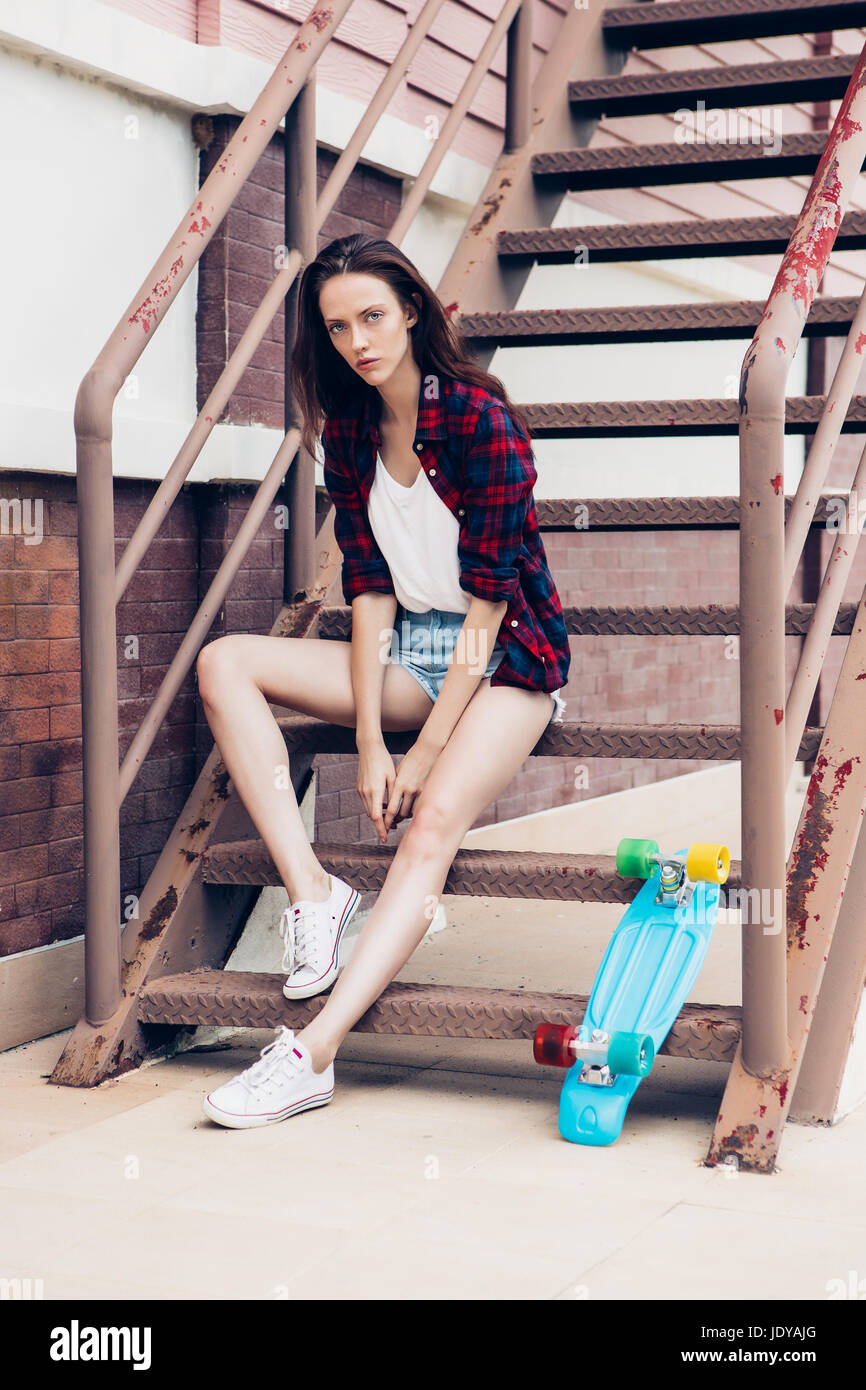 Belle jeune fille en chemise à carreaux et jeans taille basse short shorts  et baskets blanches s'asseoir sur l'escalier avec son mini bleu skateboard  longboard. U Photo Stock - Alamy