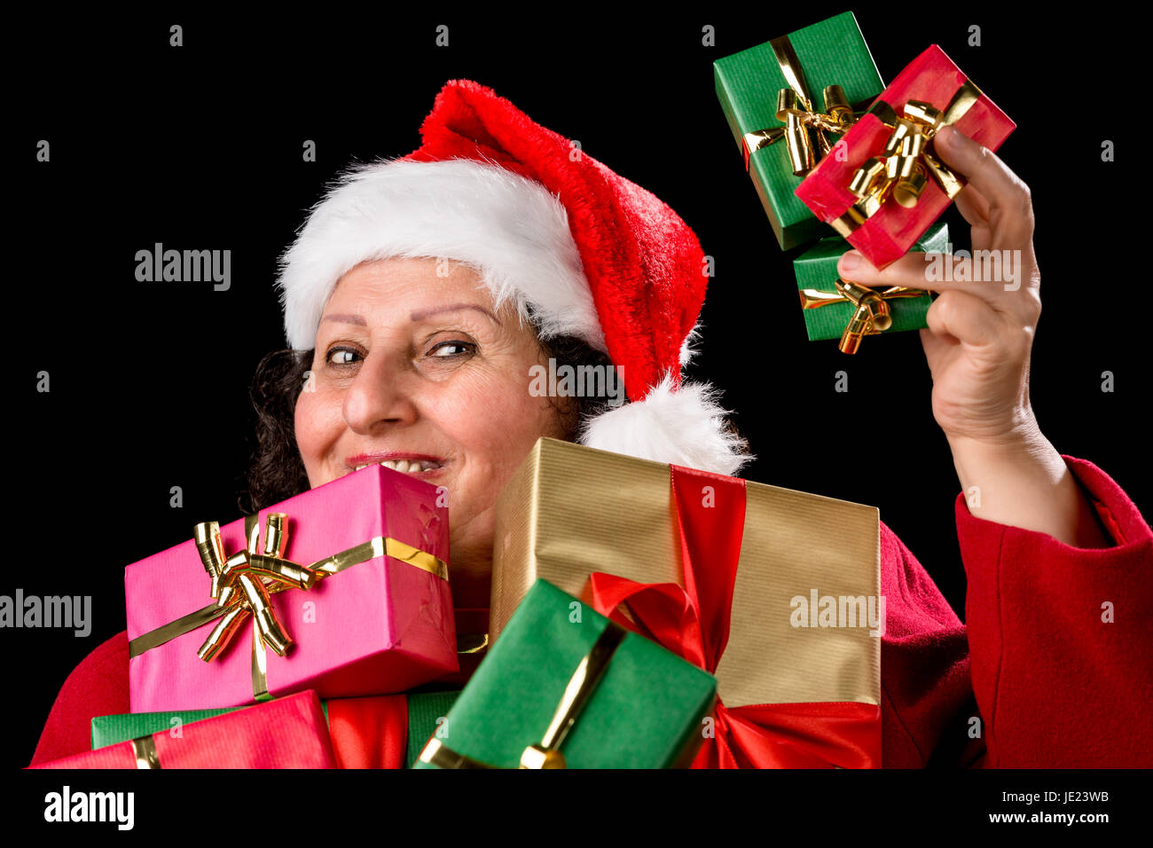 Cheerful man with Santa Claus cap offre trois petits cadeaux de Noël dans sa main gauche posée. Plus gifts en or, vert et rouge sont tenus près de sa poitrine. Isolé sur le noir. Banque D'Images