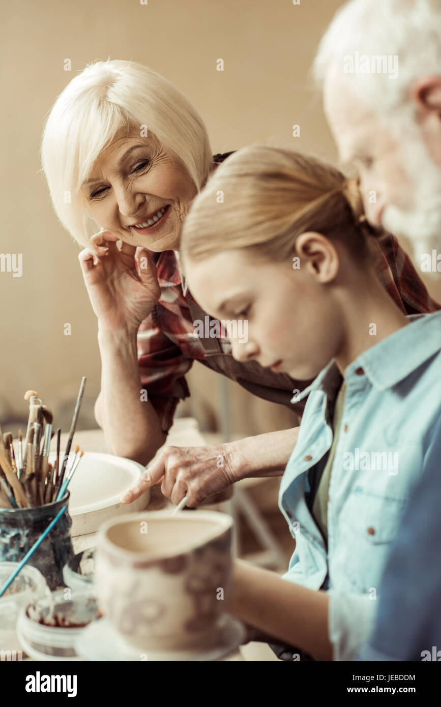 Vue latérale du girl painting pot en argile et les grands-parents d'aider à l'atelier Banque D'Images