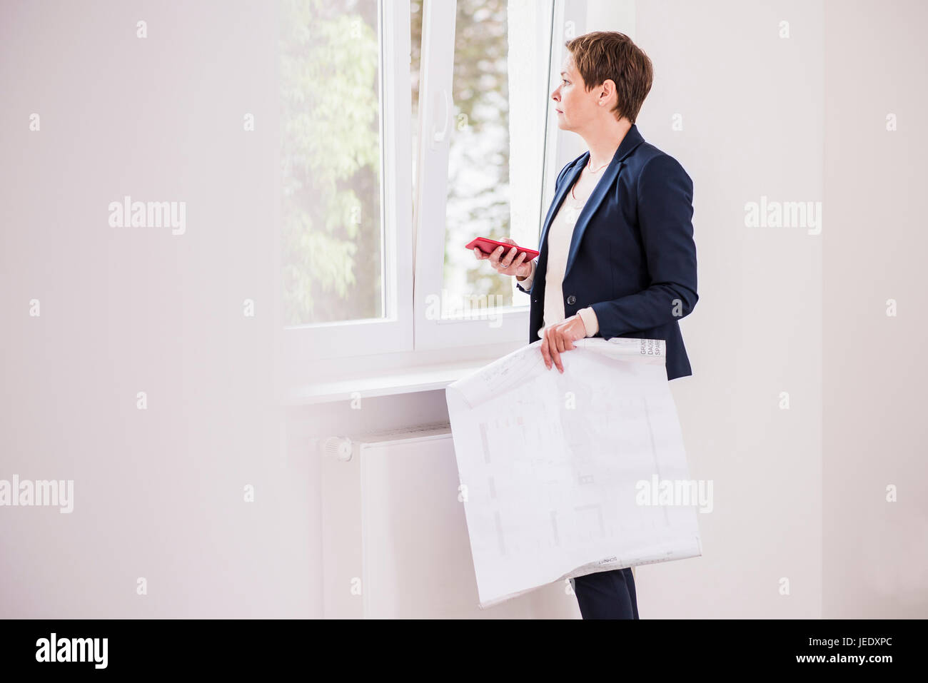 Femme dans nouvel appartement avec plan et téléphone cellulaire hors de la fenêtre Banque D'Images