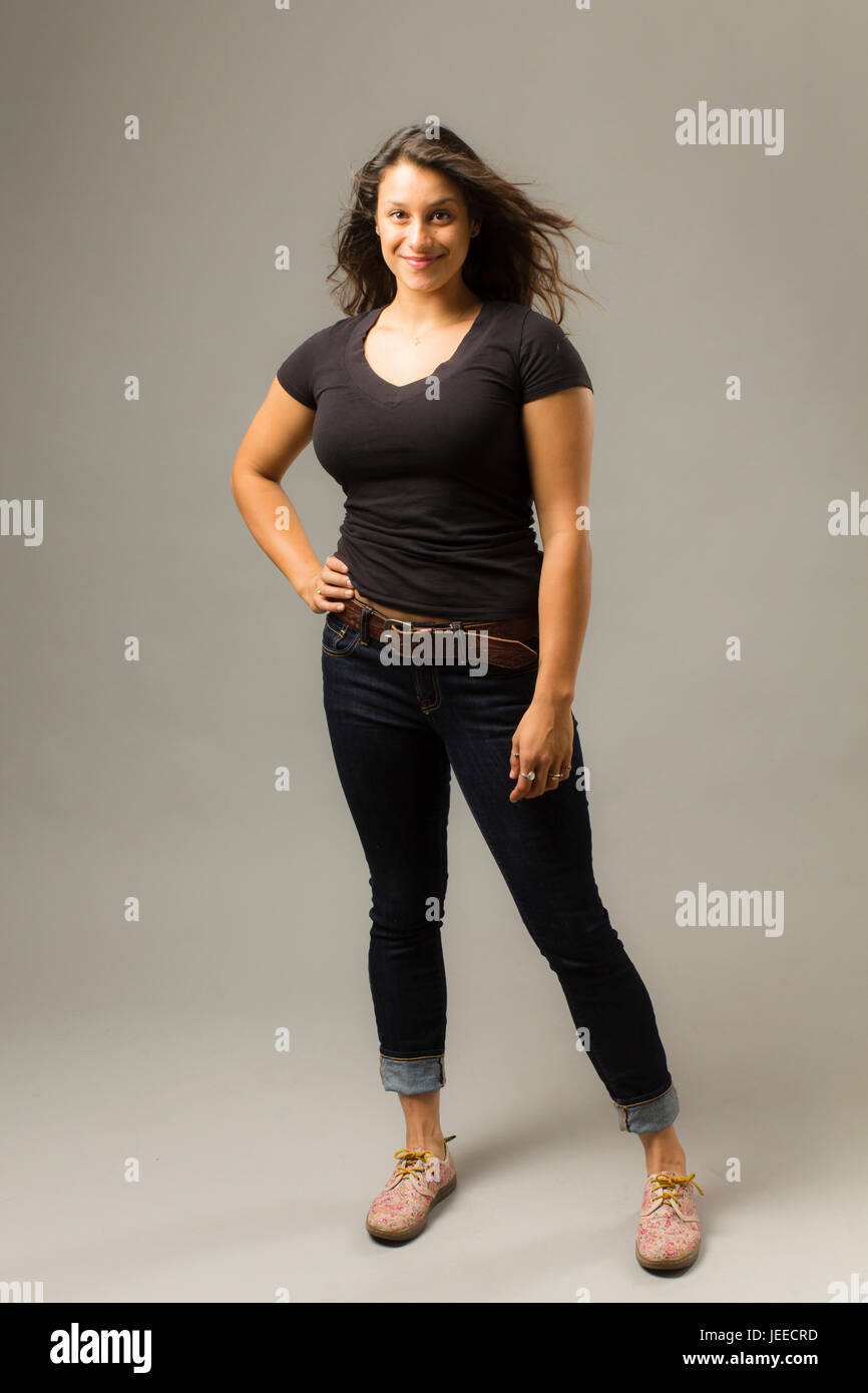 Young mixed race woman wearing a black t-shirt et jeans se tient avec les cheveux et la main sur la hanche tandis que smiling at viewer Banque D'Images