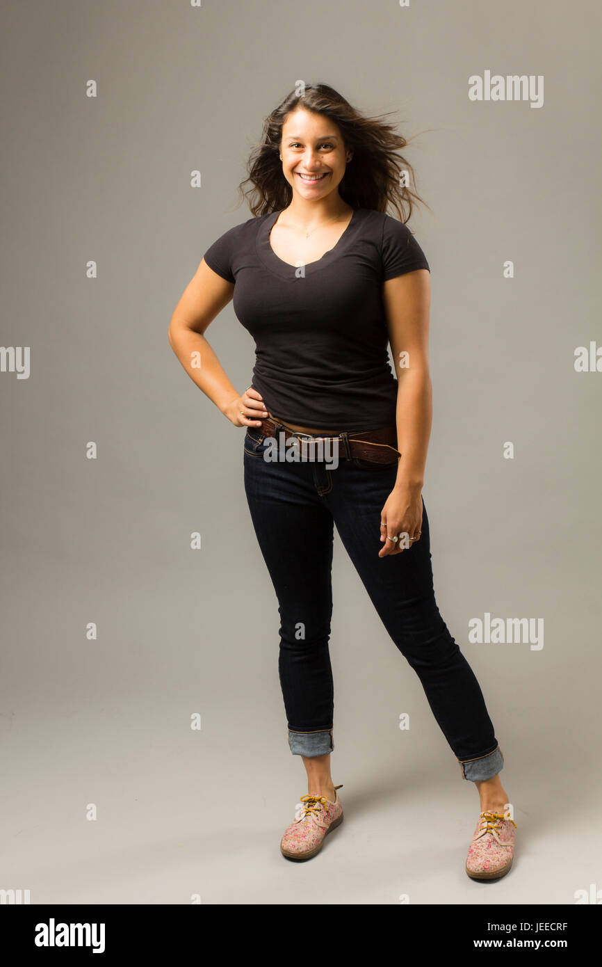 Young mixed race woman wearing a black t-shirt et jeans se tient avec les cheveux et la main sur la hanche tandis que smiling at viewer Banque D'Images