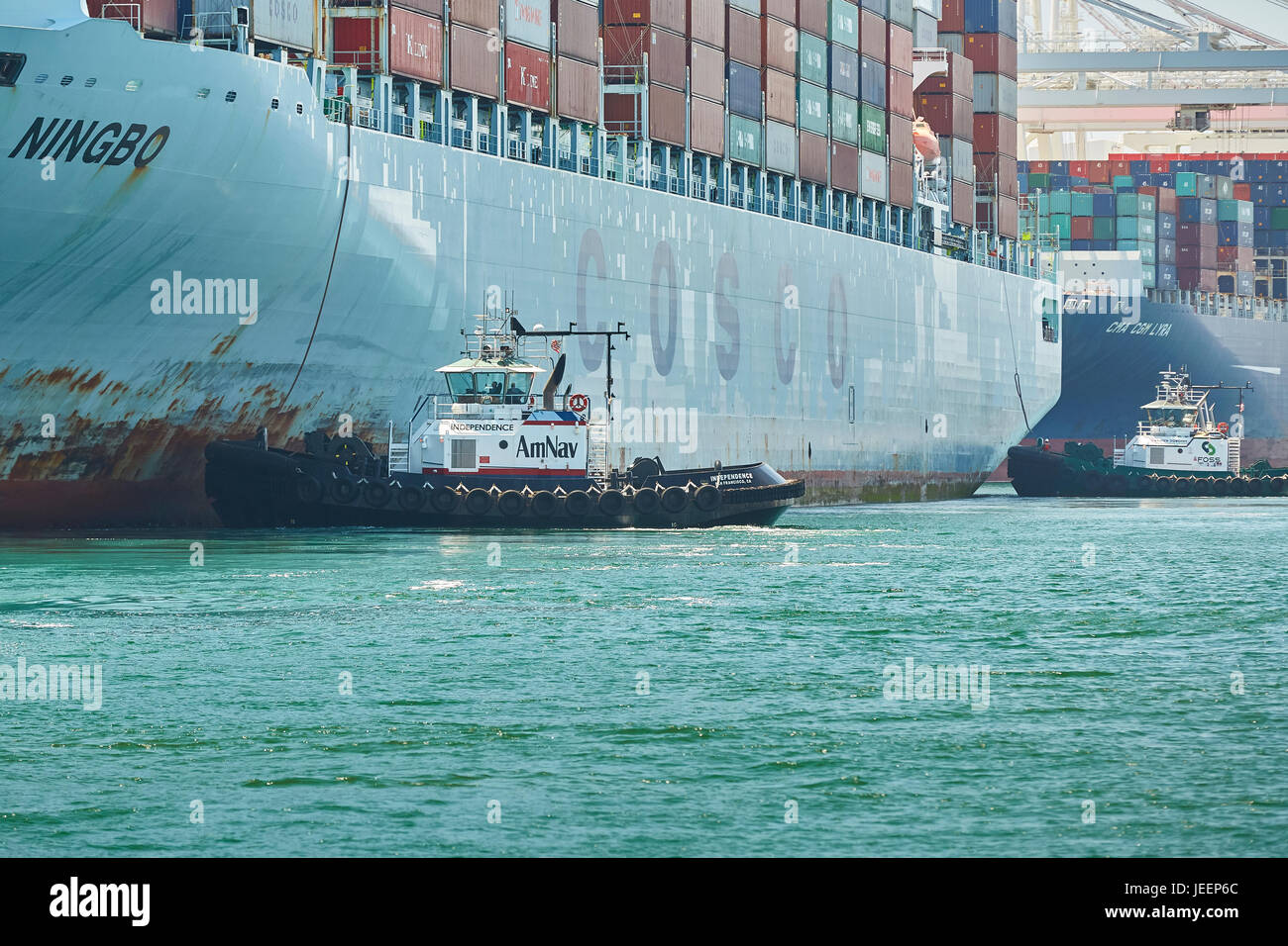 Le porte-conteneurs COSCO Géant Ningbo est guidée dans Quai J270 sur Pier J par une paire de tracteur tire sur le terminal à conteneurs de Long Beach, à Los Angeles. Banque D'Images