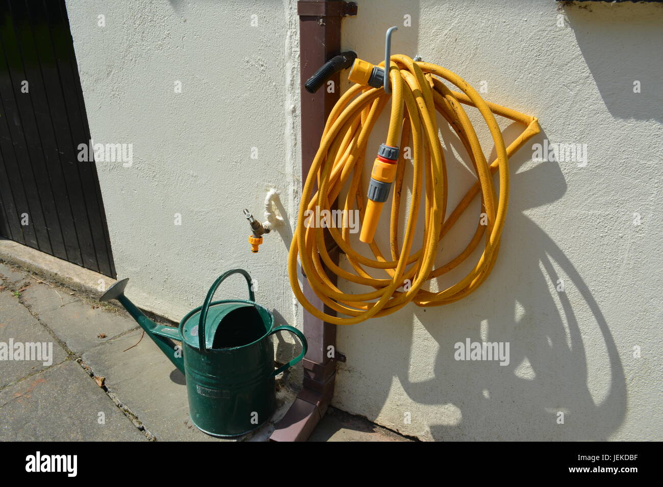 Tuyau d'arrosage jaune hanging off crochet métallique recouvert de plastique peint à l'extérieur du garage avec un arrosoir métal vert sur slabbed patio sous eau froide Banque D'Images