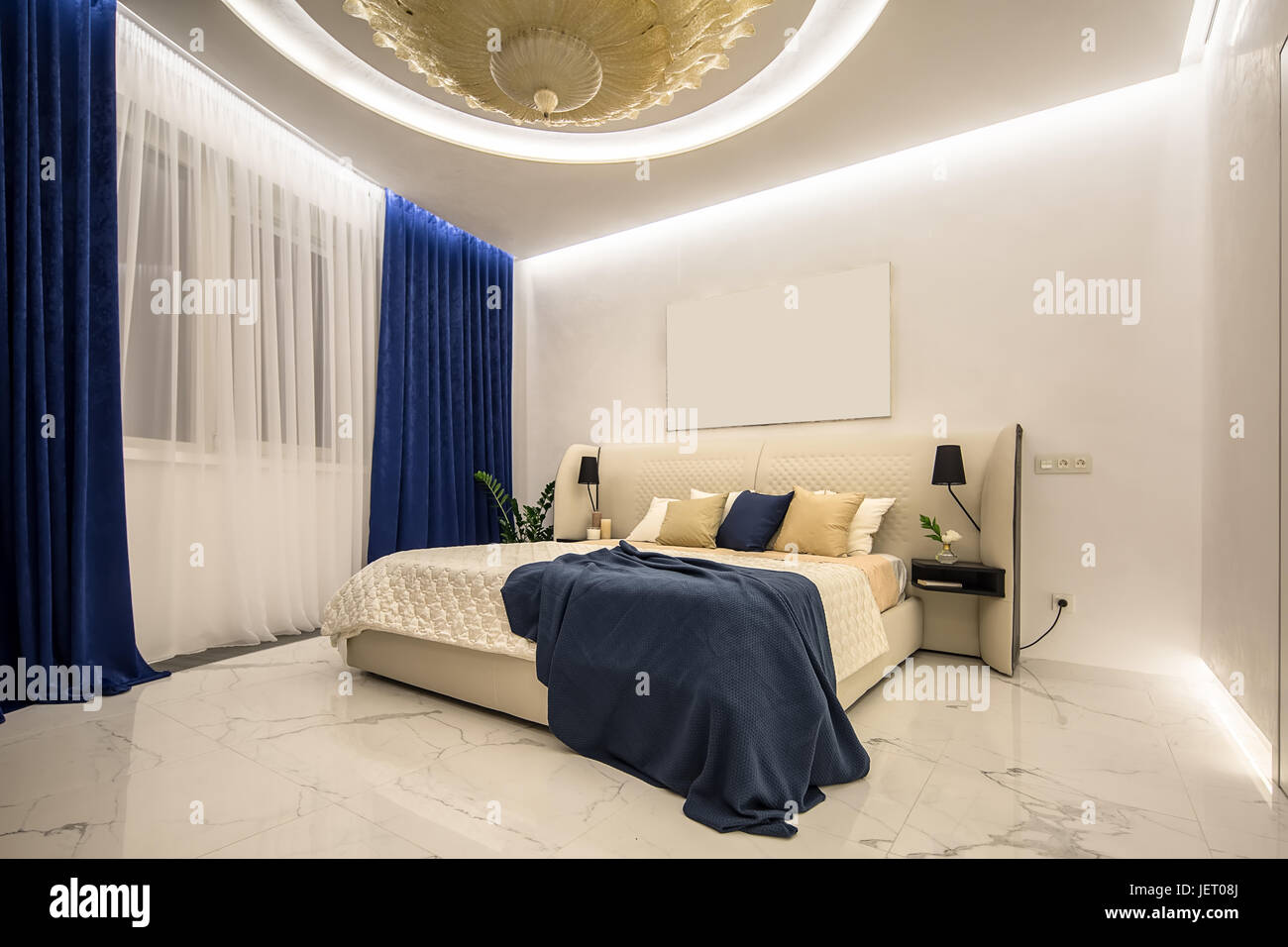 Chambre à coucher moderne éclairé par un grand lustre doré fantaisie sur le  plafond. Il y a un lit de couleur beige avec des oreillers multicolore et  d'un plaid bleu, étagères Photo