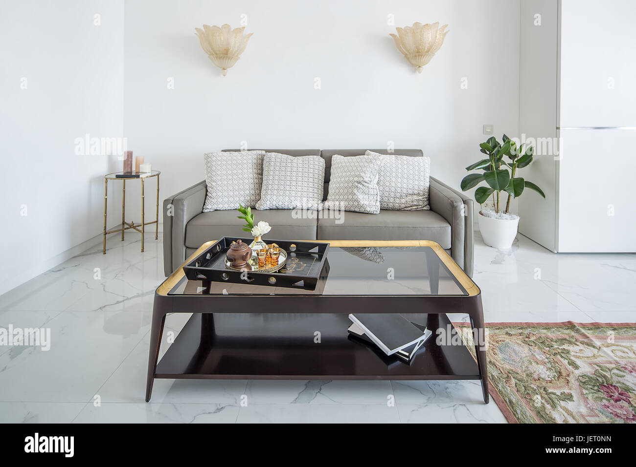 Intérieur moderne blanc avec carrelage au sol avec un tapis. Il y a des lampes dorées fantaisie sur le mur gris, canapé avec coussins, table avec une table en verre wi Banque D'Images