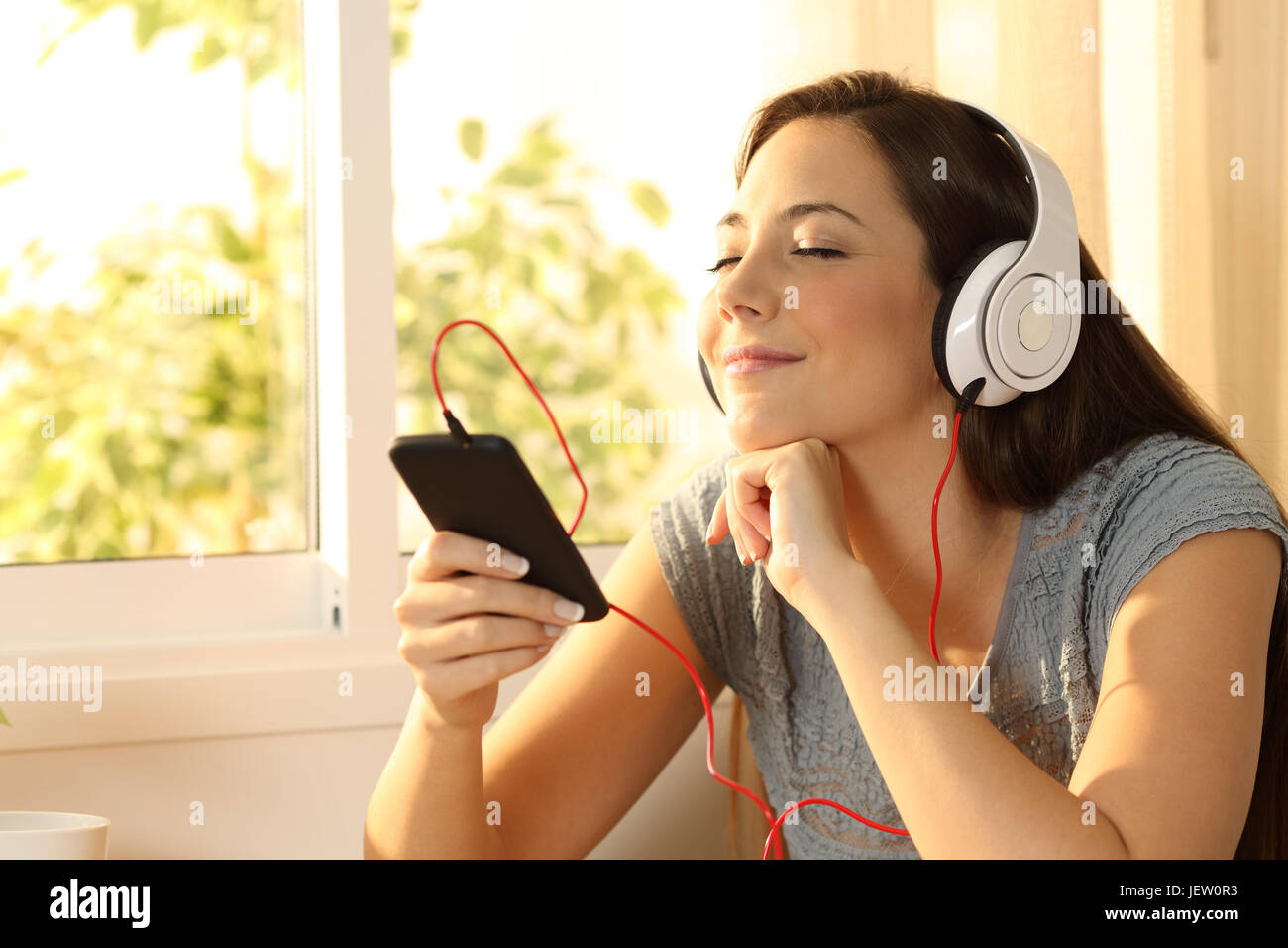 Relaxed woman listening music avec un casque et un téléphone intelligent à la maison avec une fenêtre à l'arrière-plan Banque D'Images