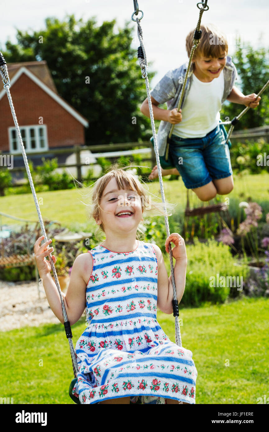 Smiling boy wearing shirt et short en jean et une fille dans une robe d'été sur des balançoires dans un jardin. Banque D'Images