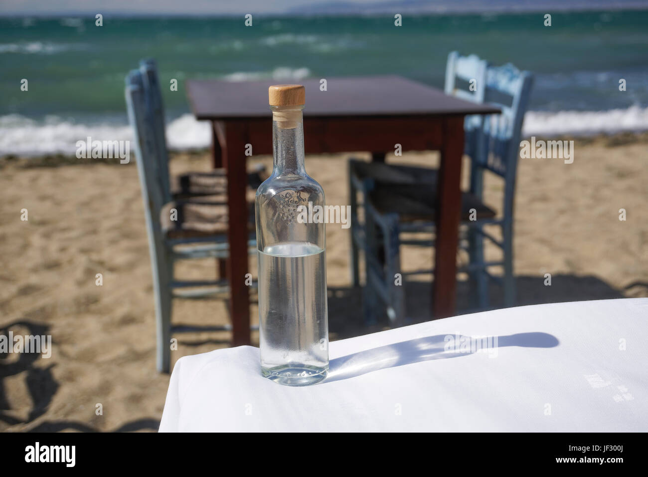 Le flacon en verre d'Ouzo Tsipouro, verre par plage grecque taverne floue avec table et chaises en bois fond bleu. Banque D'Images