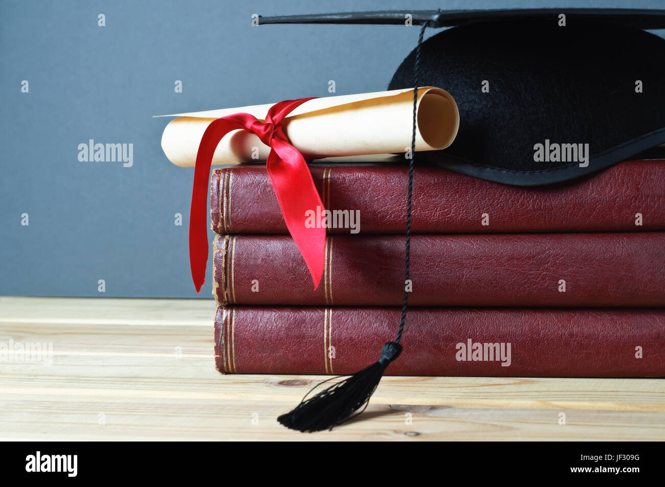 L'obtention du diplôme mortier et faites défiler jusqu'à égalité avec ruban rouge en haut d'une pile de vieux livres usés, sur une table en bois clair. Fond gris. Banque D'Images