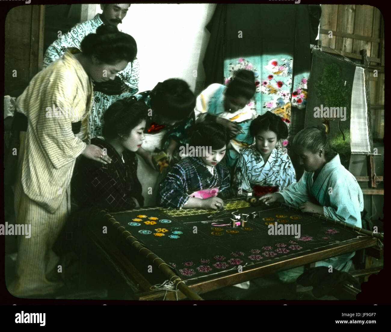 Une photographie vintage de la période Meiji montrant un groupe de femmes et un homme regardant une jeune femme travailler, reflétant les coutumes sociales japonaises Banque D'Images
