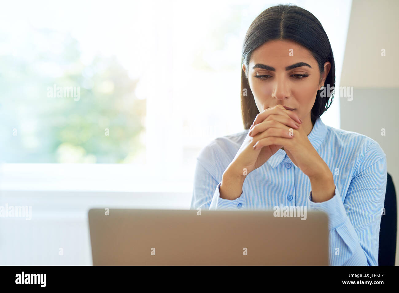 Young businesswoman sérieuse avec une expression inquiète en train de lire l'information sur son ordinateur portable avec les mains jointes contre une fenêtre haute avec spa de copie Banque D'Images