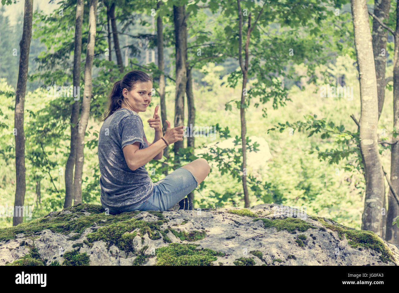 Jeune femme recherche à travers son épaule sur un rocher de la forêt. Tir fusil imaginaire les doigts. Banque D'Images