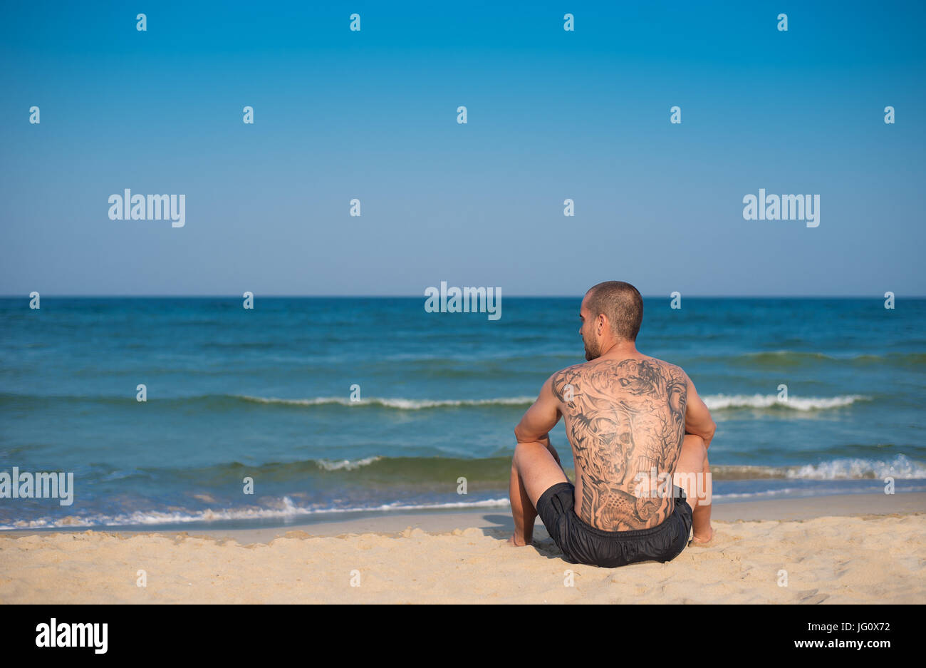 Un jeune homme avec un grand tatouage sur le dos, assis sur la plage avec vue sur la mer. Copie de l'espace sur la gauche Banque D'Images