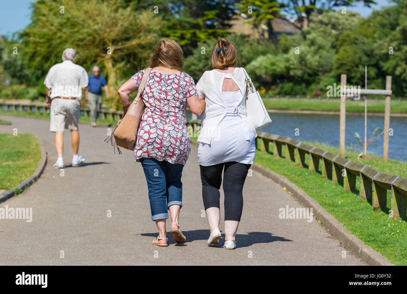 Amis féminins reliant les bras tout en marchant à travers un parc au bord d'un lac par une chaude journée d'été. Banque D'Images