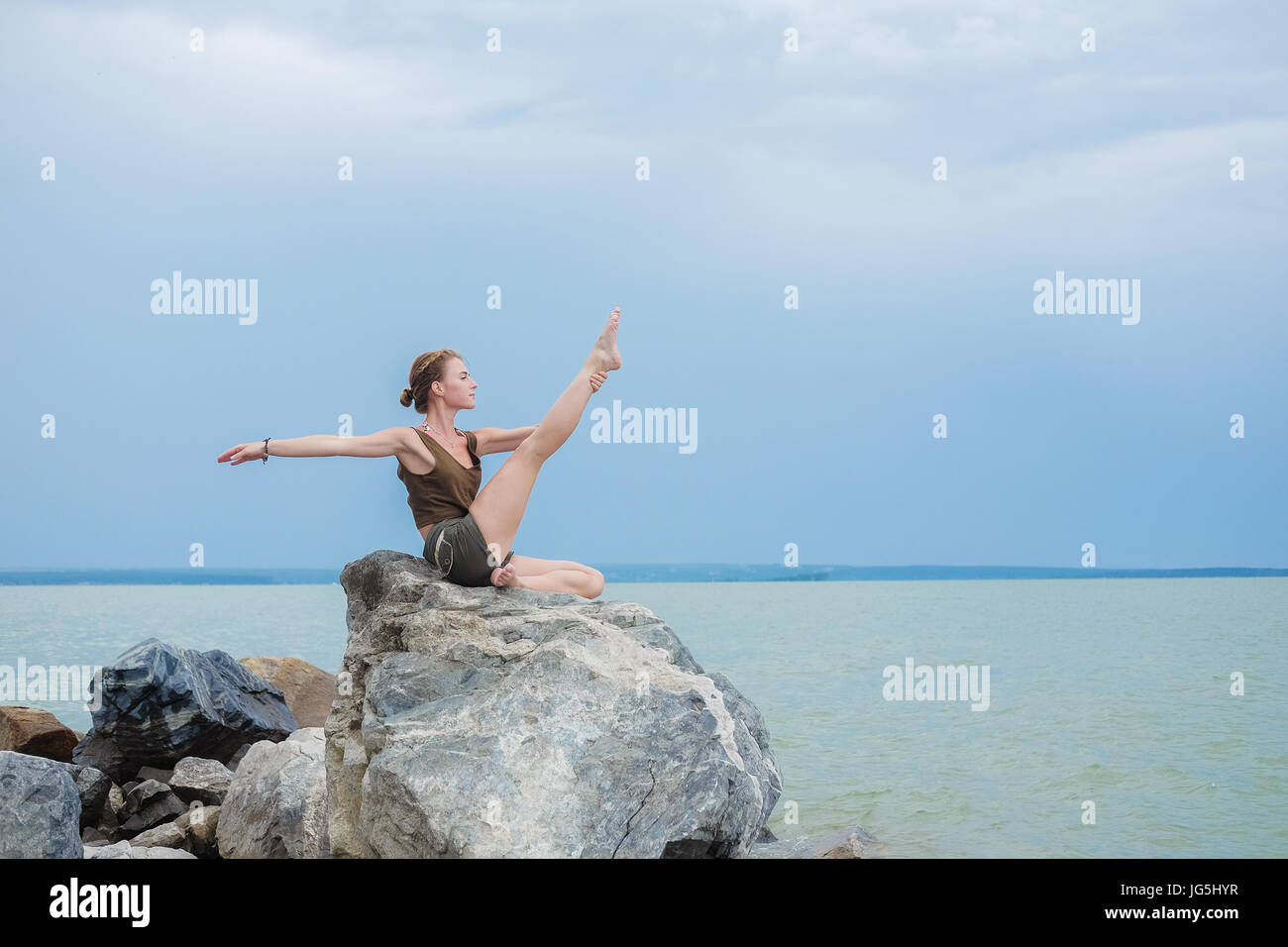 Girl practicing yoga on the rocks contre le ciel bleu et la mer d'azur. Relaxation et étirement. Jeune femme sportive slim faire du yoga à l'extérieur. Banque D'Images