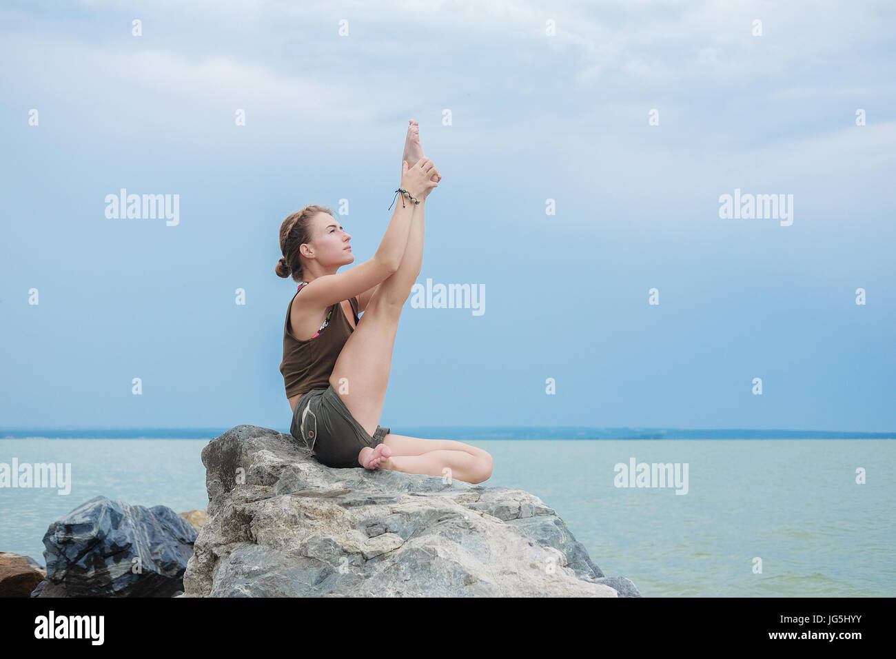Girl practicing yoga on the rocks contre le ciel bleu et la mer d'azur. Relaxation et étirement. Jeune femme sportive slim faire du yoga à l'extérieur. Banque D'Images
