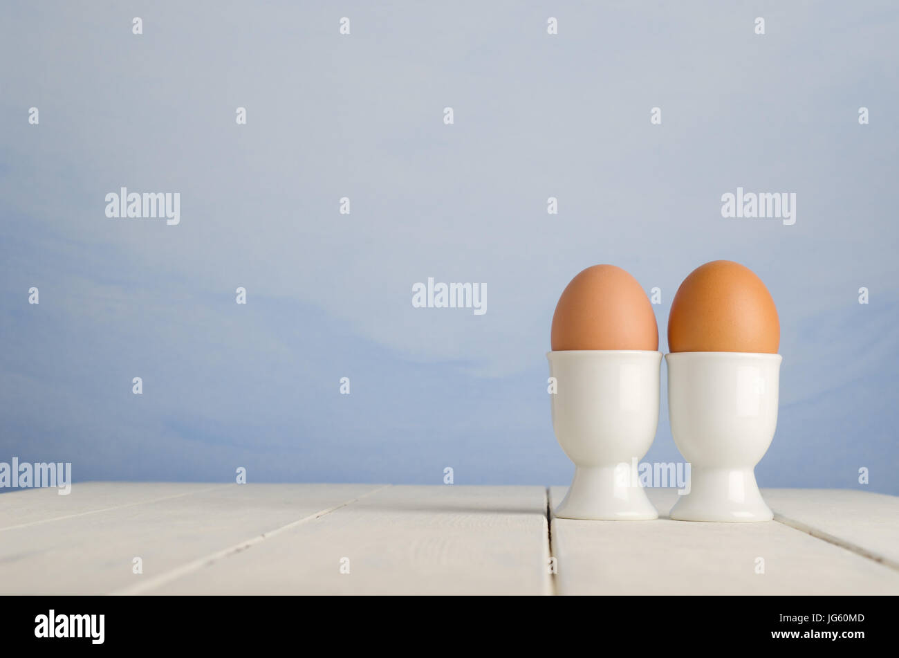Deux œufs frais brown à egg cups sur une vieille planche en bois peint crème, table de ferme, avec ciel bleu peint fond d'effet. Banque D'Images