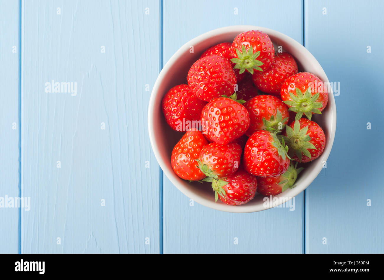 Passage tiré d'un bol blanc chine rouge contenant des fraises avec des feuilles intactes, positionné sur le côté droit d'un bleu clair bois revêtus de table. Banque D'Images