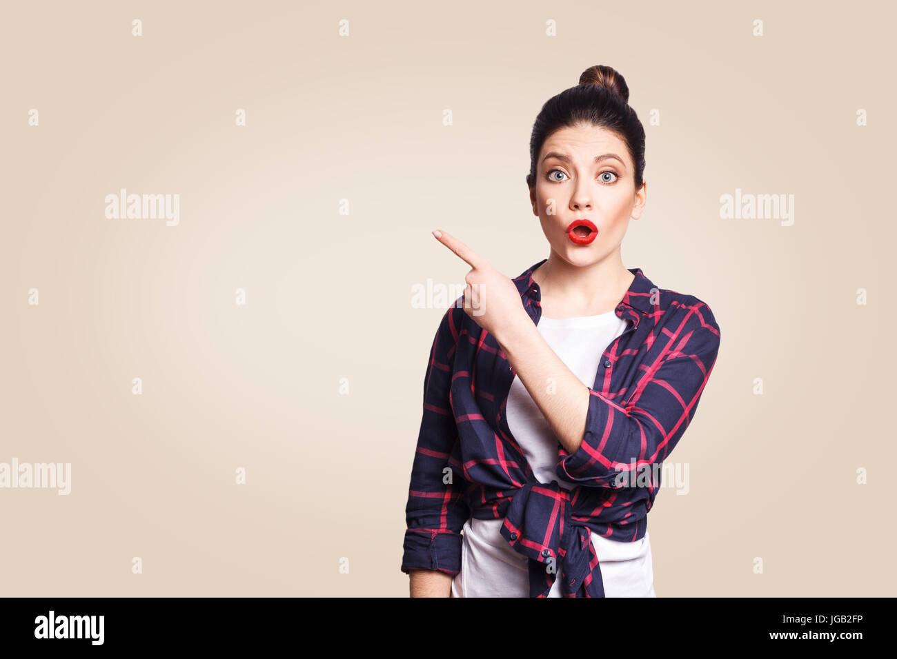 Jeune fille avec surprise et style décontracté cheveux chignon pointant son doigt sur le côté, montrant quelque chose sur le mur blanc beige avec copie espace pour votre Banque D'Images