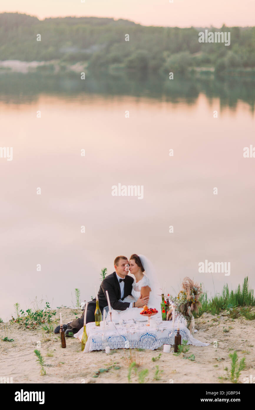 Belle photo de les mariés assis tête-à-tête tout en ayant le mariage pique-nique à l'arrière-plan de la mer au coucher du soleil. Banque D'Images