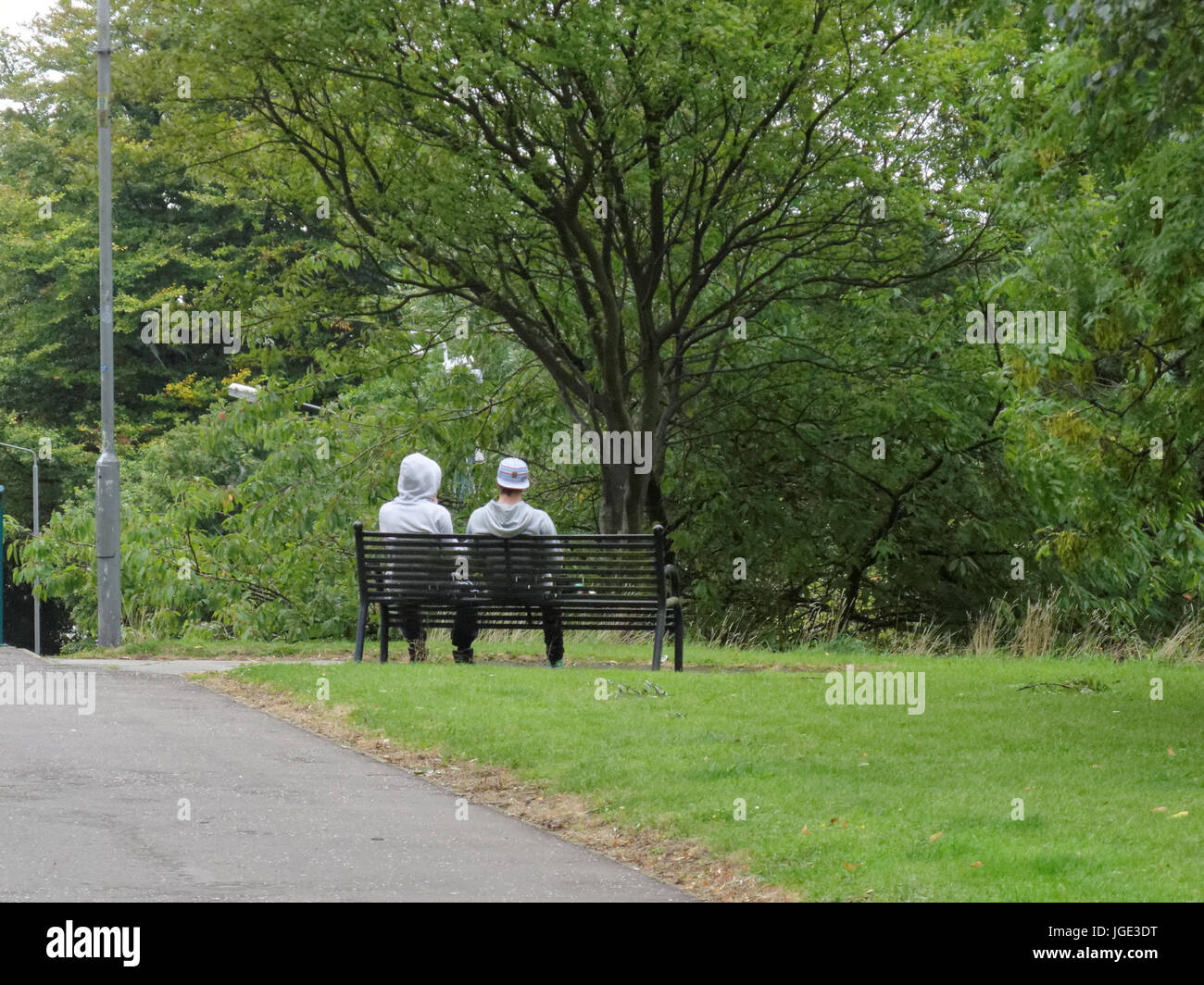 Deux hommes hommes garçons assis seuls sur un banc de parc pensivement en regardant les arbres Banque D'Images