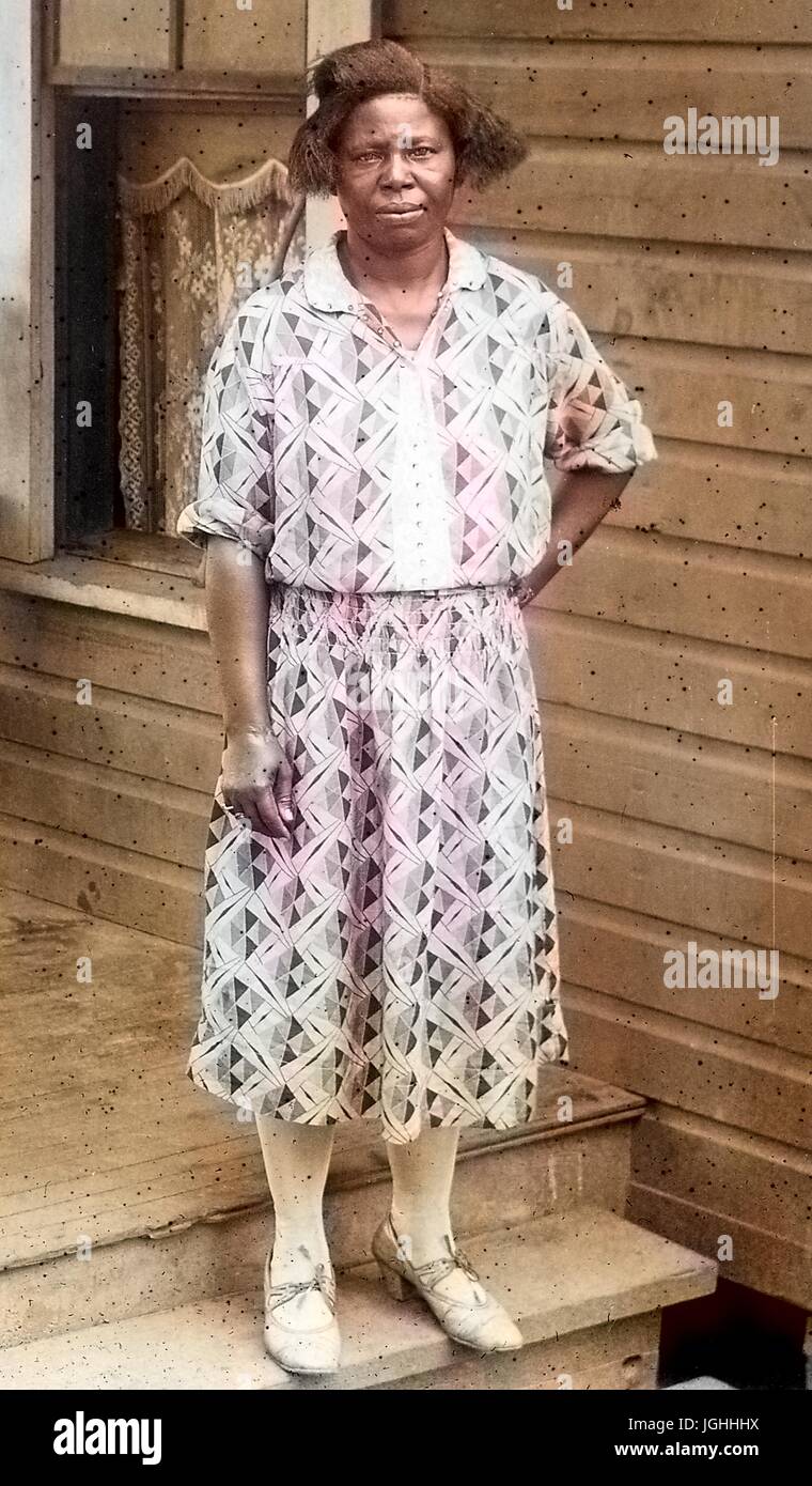 Full Length portrait of African American Woman, debout sur un porche, une main sur la hanche, portant une robe à motifs, avec une expression du visage grave, 1930. Remarque : l'image a été colorisée numériquement à l'aide d'un processus moderne. Les couleurs peuvent ne pas être exacts à l'autre. Banque D'Images